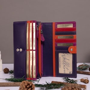 Puede incluir: Una cartera de cuero multicolor en tonos morado, naranja y rojo. La cartera está abierta, revelando múltiples ranuras para tarjetas, una ventana transparente para identificación y un compartimento con cremallera con detalles dorados.