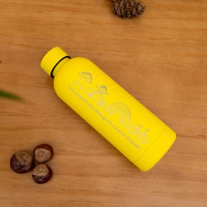 Könnte beinhalten: Eine gelbe Wasserflasche mit schwarzem Deckel und silbernem Design. Die Flasche hat einen Regenbogen und den Text "Ich habe eine schöne Botschaft für eine positive Einstellung". Die Flasche liegt auf einer Holzoberfläche.