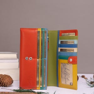 May include: A multi-coloured leather wallet featuring a vibrant orange exterior with two silver snap buttons. The interior displays a lime green lining, yellow card slots, and blue accents, with several gold-toned cards inserted into the slots.