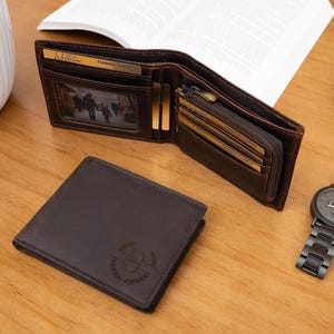 Puede incluir: Una cartera de cuero marrón, abierta para mostrar ranuras para tarjetas y un compartimento con cremallera, junto a una cartera cerrada y un reloj de pulsera negro. La cartera abierta tiene una foto familiar. El reloj tiene una correa de metal negro.