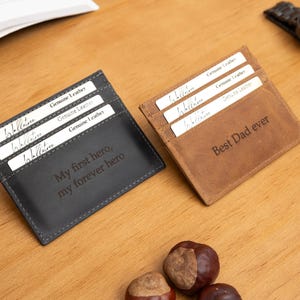 Op de afbeelding: Twee leren kaarthouders, één zwart en één bruin, met de tekst "My first hero, my forever hero" en "Best Dad ever". Elk bevat kaarten met het label "Genuine Leather". Een horloge en een open boek op de achtergrond.