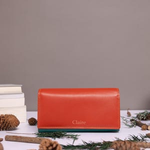 May include: A rectangular orange wallet with a dark green base and the name Claire embossed in gold lettering on the front. The wallet features a smooth finish and a structured design, positioned horizontally on a flat surface.