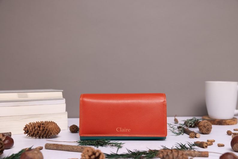 May include: A rectangular orange wallet with a dark green base and the name Claire embossed in gold lettering on the front. The wallet features a smooth finish and a structured design, positioned horizontally on a flat surface.