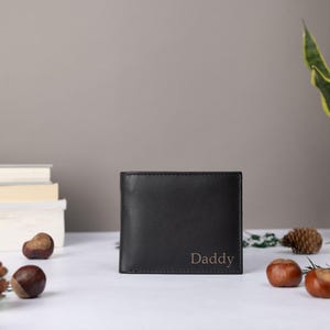 May include: A black bifold wallet featuring the word Daddy embossed in a simple serif font on the lower right corner. The wallet is constructed from a smooth, dark material and maintains a rectangular shape with clean, straight edges.