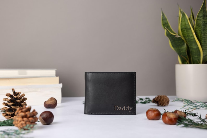 May include: A black bifold wallet featuring the word Daddy embossed in a simple serif font on the lower right corner. The wallet is constructed from a smooth, dark material and maintains a rectangular shape with clean, straight edges.