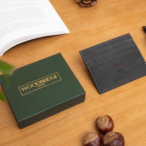 May include: A dark green gift box with gold lettering reading "WOODBRIDGE LONDON" sits next to a black leather card holder with "Dad" embossed. A wooden watch and open book are also on the wooden surface.