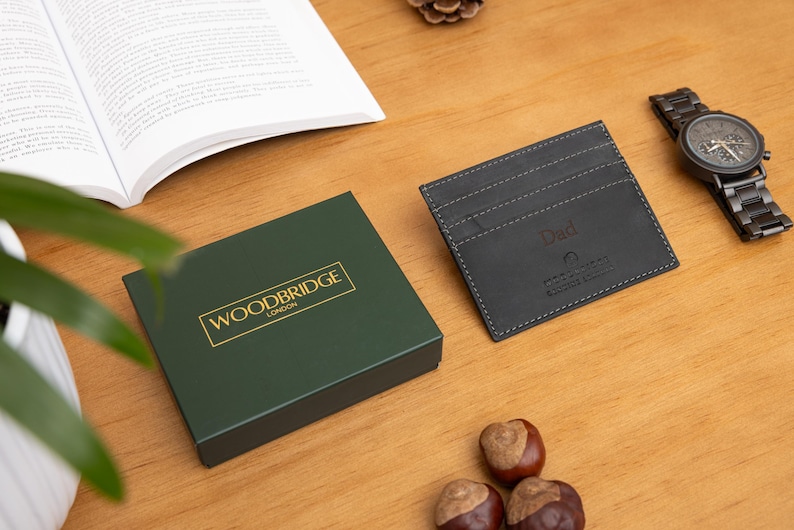 May include: A dark green gift box with gold lettering reading "WOODBRIDGE LONDON" sits next to a black leather card holder with "Dad" embossed. A wooden watch and open book are also on the wooden surface.