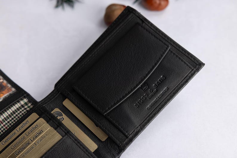 May include: A black leather wallet featuring multiple card slots, a coin pocket with a flap, and the embossed text BIGGS & BATE LONDON GENUINE LEATHER. The interior includes several gold-colored cards labeled Genuine Leather and a plaid fabric lining.
