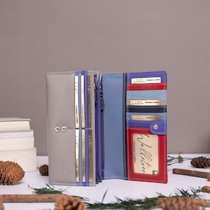May include: A multi-colored leather wallet featuring a grey exterior and an interior with blue, red, and grey card slots. The wallet includes multiple vertical card pockets, a clear identification window, and a central zippered compartment.