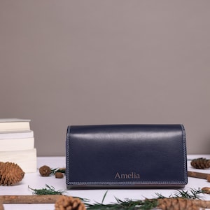 May include: A rectangular navy blue leather wallet featuring a smooth finish and visible stitching along the edges. The name Amelia is embossed in a simple font on the front lower center of the wallet, which is positioned horizontally.