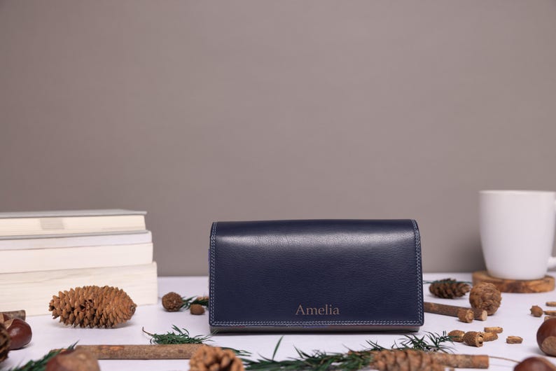 May include: A rectangular navy blue leather wallet featuring a smooth finish and visible stitching along the edges. The name Amelia is embossed in a simple font on the front lower center of the wallet, which is positioned horizontally.