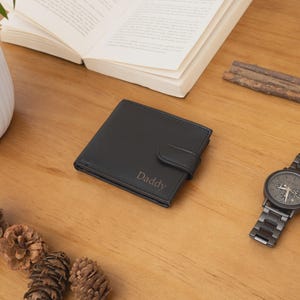 May include: A black leather wallet with the word "Daddy" embossed on the front, next to a dark metal watch, open book, and pine cones on a wooden surface. A white potted plant is in the background.