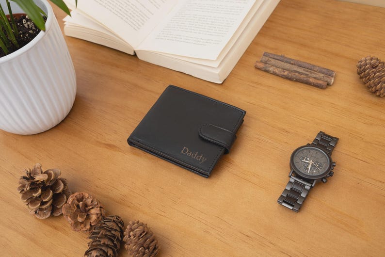 May include: A black leather wallet with the word "Daddy" embossed on the front, next to a dark metal watch, open book, and pine cones on a wooden surface. A white potted plant is in the background.