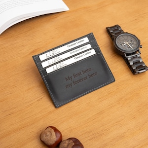 May include: A dark gray leather cardholder with the text "My first hero, my forever hero" is displayed on a wooden surface. Three cards are visible in the cardholder. A black watch with a metal band is next to the cardholder.