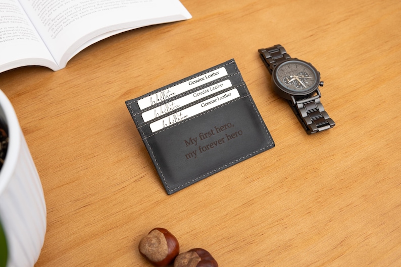 May include: A dark gray leather cardholder with the text "My first hero, my forever hero" is displayed on a wooden surface. Three cards are visible in the cardholder. A black watch with a metal band is next to the cardholder.