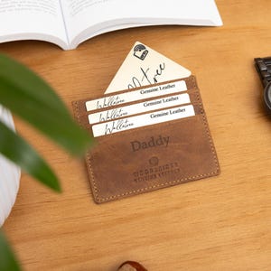 May include: A brown leather wallet with the word "Daddy" embossed on the front, holding several white cards. A black wristwatch with a dark face and band sits to the right. The items are on a wooden surface.