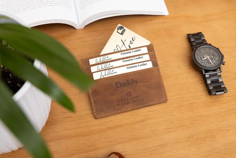 May include: A brown leather wallet with the word "Daddy" embossed on the front, holding several white cards. A black wristwatch with a dark face and band sits to the right. The items are on a wooden surface.
