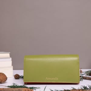 May include: A rectangular olive green wallet with a smooth finish and a flap closure. The name Amanda is embossed in a light font on the lower center of the front flap. The wallet has a slim profile and a solid, uniform color throughout the exterior.