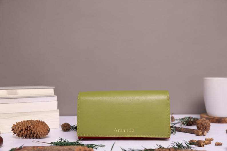 May include: A rectangular olive green wallet with a smooth finish and a flap closure. The name Amanda is embossed in a light font on the lower center of the front flap. The wallet has a slim profile and a solid, uniform color throughout the exterior.