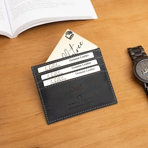 May include: A black leather cardholder with the word "Dad" embossed on the front, holding several cards. A black wristwatch with a metal band sits to the right. The cardholder has the words "Genuine Leather" on the cards.