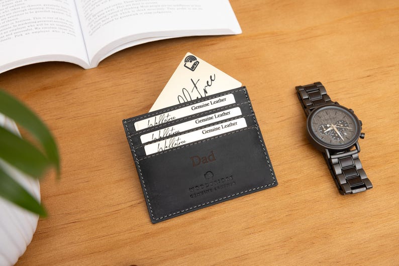 May include: A black leather cardholder with the word "Dad" embossed on the front, holding several cards. A black wristwatch with a metal band sits to the right. The cardholder has the words "Genuine Leather" on the cards.