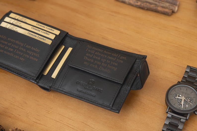 May include: Black leather wallet with engraved text and a matching black wristwatch. The wallet is open, revealing card slots and more engraved text. The watch has a dark face and a black metal band.