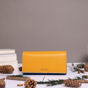 May include: A rectangular mustard yellow leather wallet featuring the name Sarah embossed in a simple serif font on the front flap. The wallet has a smooth, structured design with a visible blue interior lining along the bottom edge.