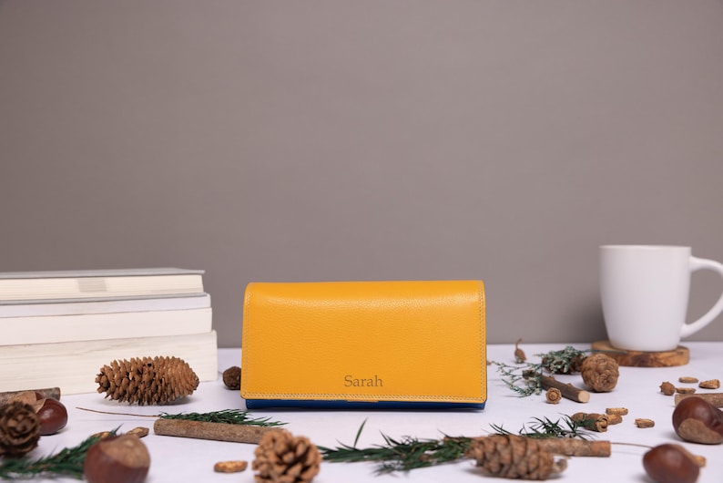 May include: A rectangular mustard yellow leather wallet featuring the name Sarah embossed in a simple serif font on the front flap. The wallet has a smooth, structured design with a visible blue interior lining along the bottom edge.