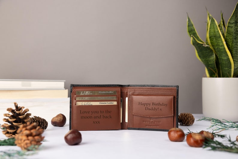 May include: A bi-fold wallet made of black and brown leather, featuring an open interior with card slots and a coin pocket. The interior is engraved with the text 'Love you to the moon and back XXX' and 'Happy Birthday Daddy! x' on the coin flap.