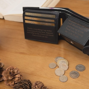 May include: Black leather wallet, open to display card slots and a bill compartment. The wallet has embossed text inside. Several coins are scattered on the wooden surface. Pinecones and a white vase are also visible.