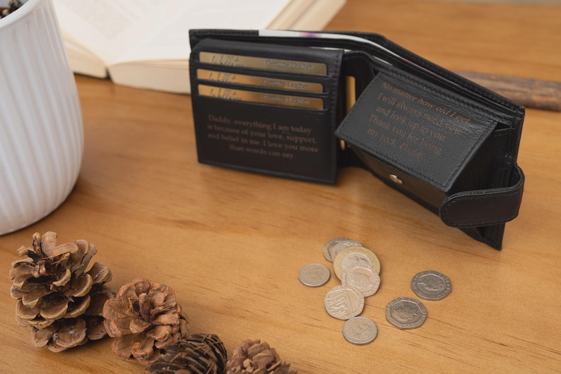 May include: Black leather wallet, open to display card slots and a bill compartment. The wallet has embossed text inside. Several coins are scattered on the wooden surface. Pinecones and a white vase are also visible.