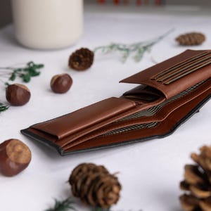 May include: A bi-fold wallet made of smooth brown leather with a black exterior edge. The interior features multiple card slots, a bill compartment, and a dark fabric lining with a repeating geometric pattern visible inside the open sections.