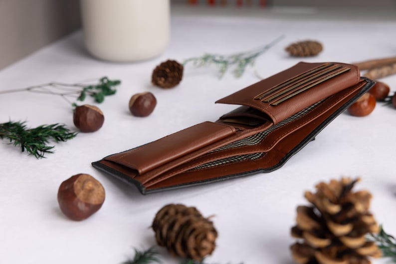 May include: A bi-fold wallet made of smooth brown leather with a black exterior edge. The interior features multiple card slots, a bill compartment, and a dark fabric lining with a repeating geometric pattern visible inside the open sections.