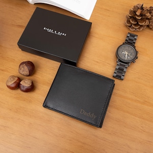 May include: A black leather bifold wallet with the word Daddy embossed on the front, accompanied by a black gift box labeled Biggs & Bane, a dark metal wristwatch with a link band, three brown chestnuts, and three pine cones.