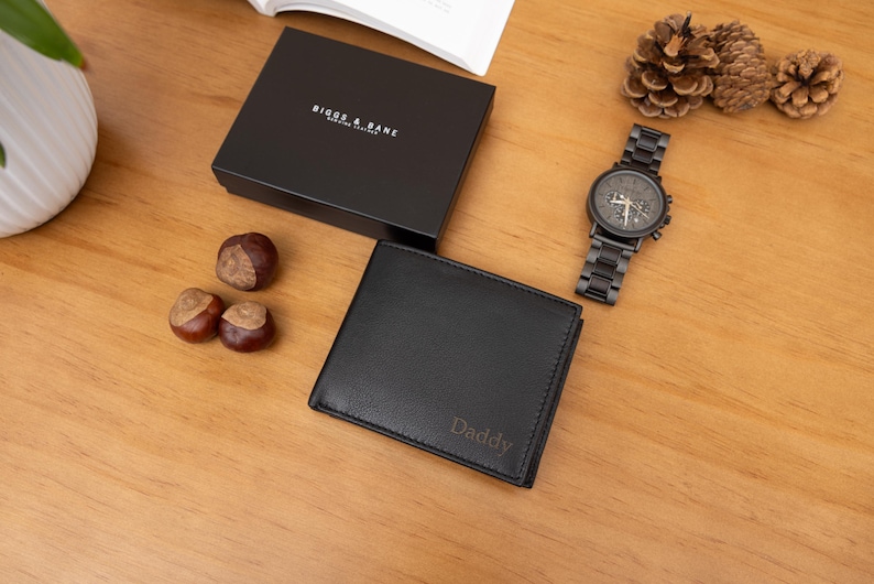 May include: A black leather bifold wallet with the word Daddy embossed on the front, accompanied by a black gift box labeled Biggs & Bane, a dark metal wristwatch with a link band, three brown chestnuts, and three pine cones.