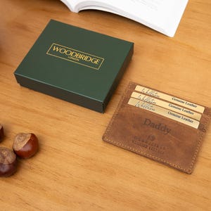 May include: A brown leather wallet with the word "Daddy" embossed on it, next to a dark green box with gold lettering. A black watch and three chestnuts are also on the wooden surface.