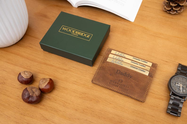 May include: A brown leather wallet with the word "Daddy" embossed on it, next to a dark green box with gold lettering. A black watch and three chestnuts are also on the wooden surface.