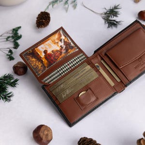 May include: A brown leather bi-fold wallet featuring a clear ID window with a printed image of a cozy indoor scene, multiple card slots, a small coin pocket, and a plaid fabric lining. The interior is embossed with text and includes gold-colored cards.