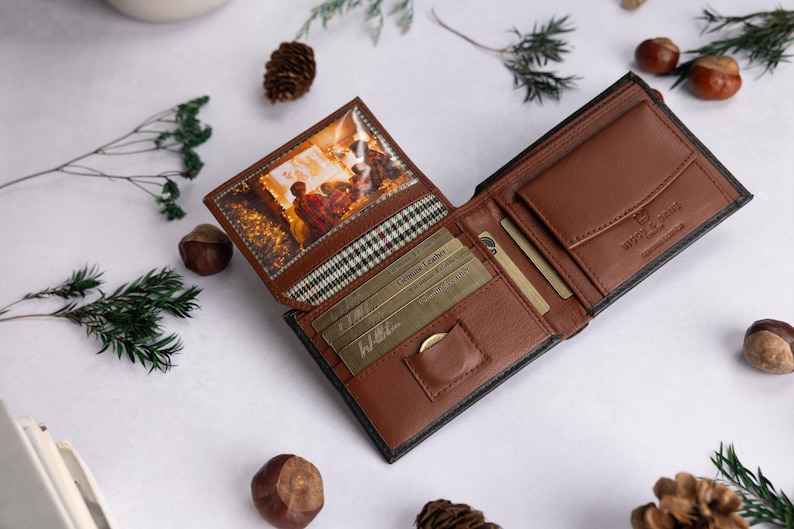 May include: A brown leather bi-fold wallet featuring a clear ID window with a printed image of a cozy indoor scene, multiple card slots, a small coin pocket, and a plaid fabric lining. The interior is embossed with text and includes gold-colored cards.