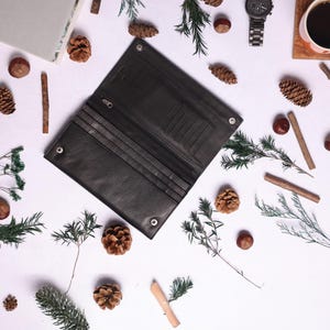 May include: A black leather wallet with multiple card slots and a billfold. The wallet is open and lying on a white surface with pine cones and other natural elements scattered around it.