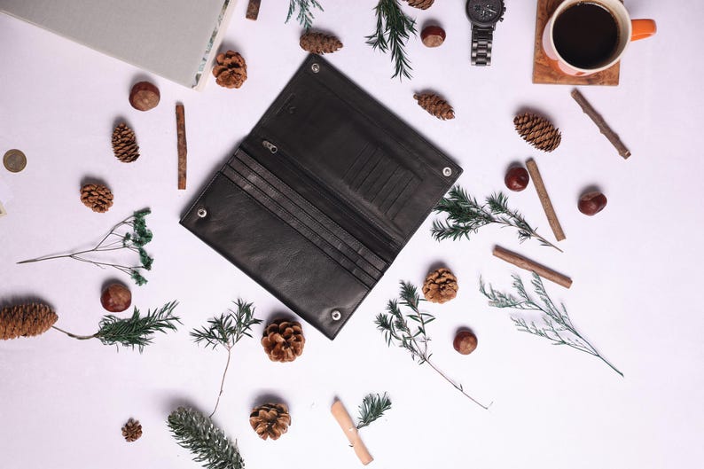 May include: A black leather wallet with multiple card slots and a billfold. The wallet is open and lying on a white surface with pine cones and other natural elements scattered around it.