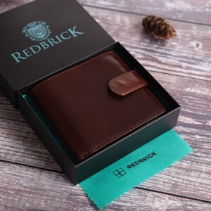May include: A brown leather wallet in a black box with the text "REDBRICK" on the box lid. The wallet has a flap closure and a small pocket on the front. The box is sitting on a wooden surface.