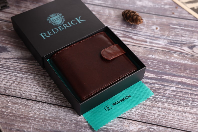 May include: A brown leather wallet in a black box with the text "REDBRICK" on the box lid. The wallet has a flap closure and a small pocket on the front. The box is sitting on a wooden surface.