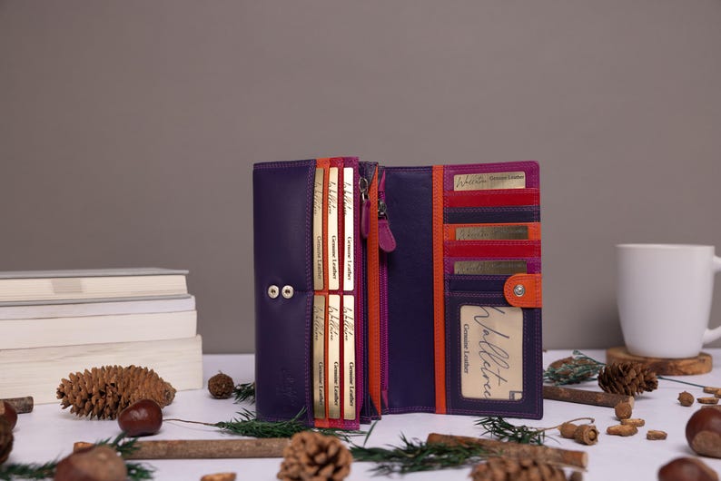 May include: A purple leather wallet with orange and pink accents. The wallet is open and has multiple card slots and a zippered compartment. The wallet is on a white surface with pine cones and twigs.