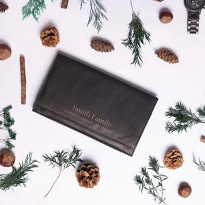 May include: A black leather wallet with the text "Smith Family" and "Celebrate life, one journey at a time" embossed in gold. The wallet is surrounded by pine cones, twigs, and greenery on a white background.