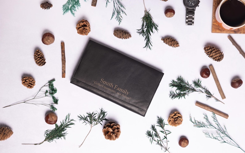 May include: A black leather wallet with the text "Smith Family" and "Celebrate life, one journey at a time" embossed in gold. The wallet is surrounded by pine cones, twigs, and greenery on a white background.