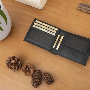 May include: Black leather wallet with multiple card slots and a bill compartment, open on a wooden surface. The wallet has text inside and is next to a black watch and pine cones. Genuine leather cards are visible.