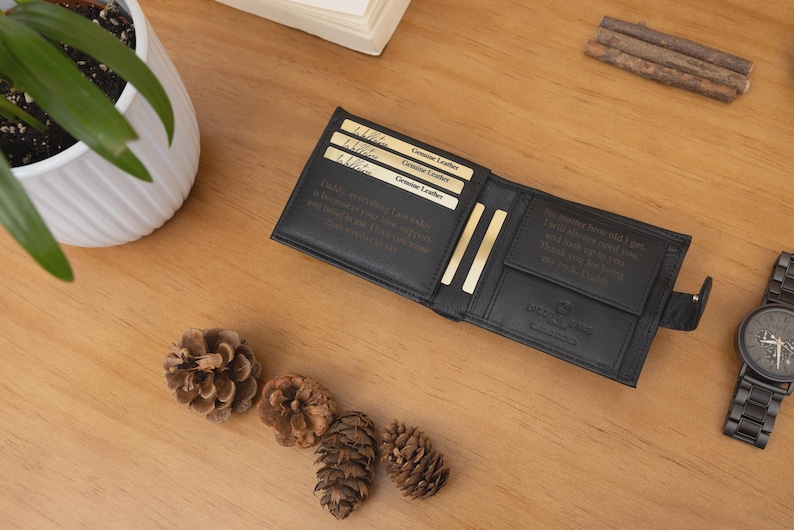 May include: Black leather wallet with multiple card slots and a bill compartment, open on a wooden surface. The wallet has text inside and is next to a black watch and pine cones. Genuine leather cards are visible.