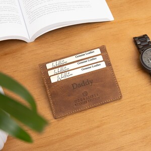 May include: A brown leather wallet with the word "Daddy" engraved on it, holding three cards. A black wristwatch with a matching metal band sits to the right. The items are on a wooden surface.