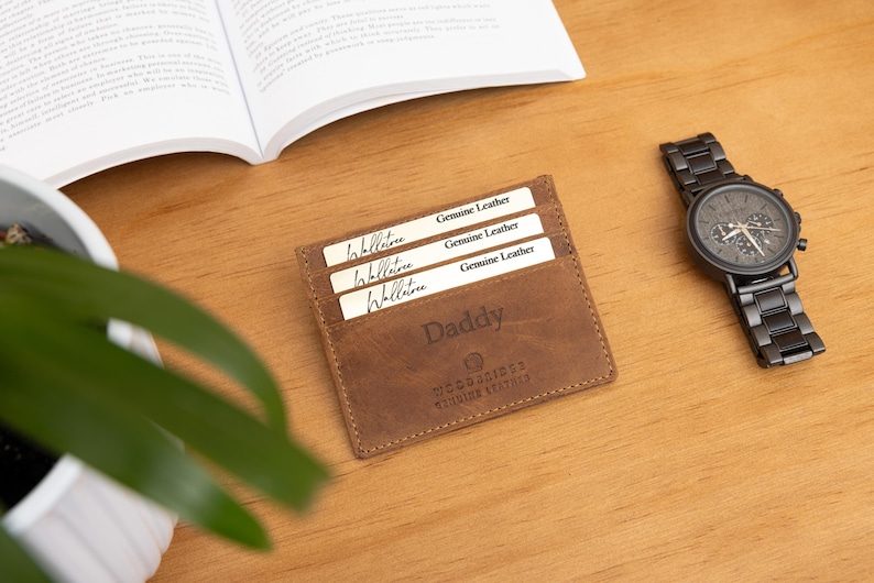 May include: A brown leather wallet with the word "Daddy" engraved on it, holding three cards. A black wristwatch with a matching metal band sits to the right. The items are on a wooden surface.